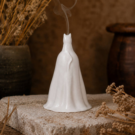 A white ceramic cloaked figure incense burner with a small stemhole and visible rising smoke, displayed on a stone slab with dried botanicals and rustic pottery in the background.