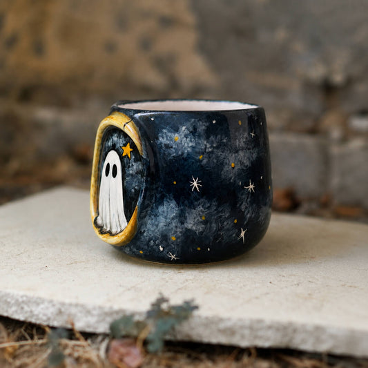 A small dark ceramic cup or candle holder with a hand-painted celestial and ghost design, resting on a stone slab outdoors.