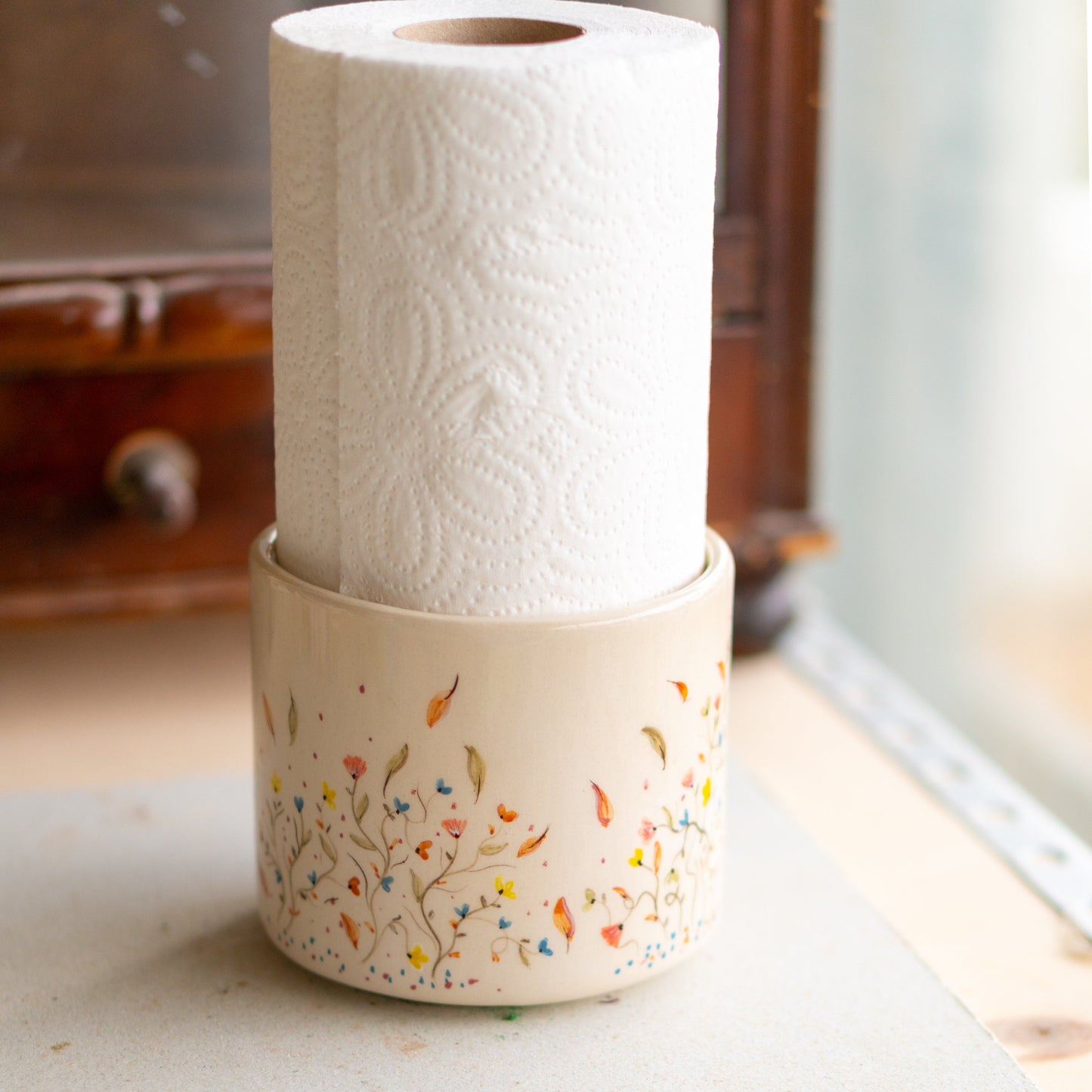 A front-facing view of a ceramic paper towel holder, illustrating the fine artistry of the painted blooms and foliage against a clean, rustic design