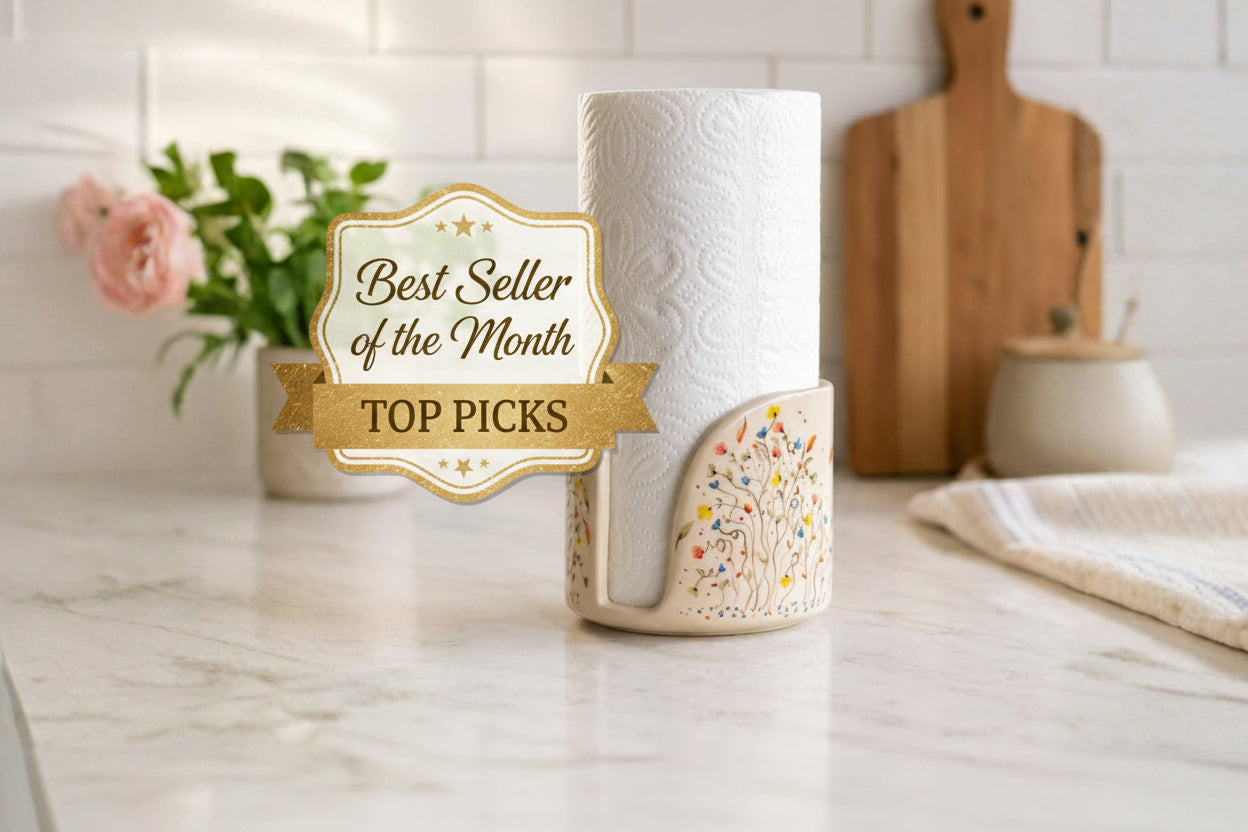 A ceramic paper towel holder with a roll of embossed paper towels, styled on a marble kitchen counter with a potted plant and wooden cutting board in the background.