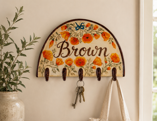 A personalized wall-mounted key rack with a cream background, orange poppy floral pattern, and the name 'Brown' in script. The rack has five dark brown hooks, one of which holds a set of keys, and another hanging hook holds a canvas tote bag.
