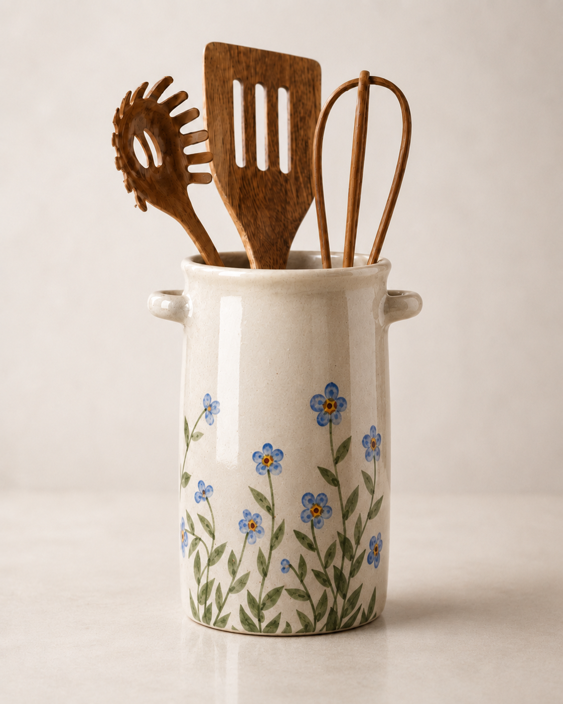 A ceramic kitchen utensil holder with side handles, decorated with a blue and green forget-me-not floral pattern. It contains three wooden kitchen utensils, including a slotted spatula, a pasta server, and a whisk, set against a clean, light beige background.