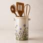 A ceramic kitchen utensil holder with a cream glaze, decorated with blue flowers and green leaves, holding wooden kitchen tools including a spatula, whisk, and spoon, set against a neutral beige background.