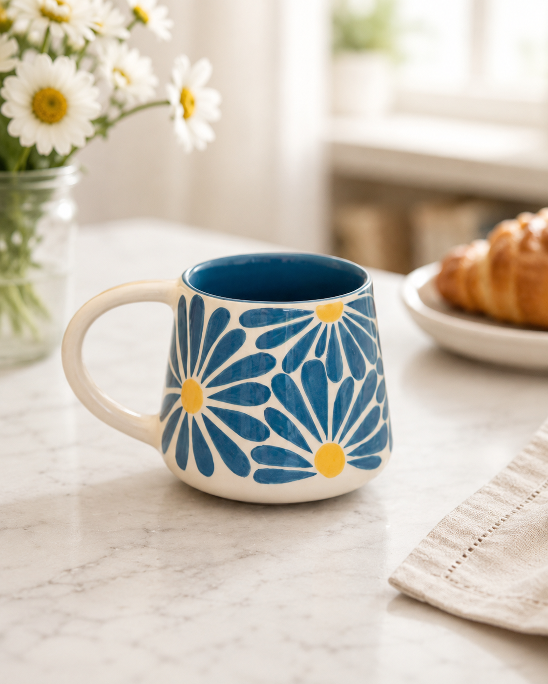 A ceramic mug with a blue and yellow daisy pattern, sitting on a marble countertop in a bright kitchen setting with flowers and a croissant blurred in the background.