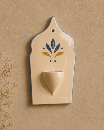 An off-white ceramic wall-mounted cup holder with a small central bowl and a decorative blue and gold floral motif, set against a textured beige wall.