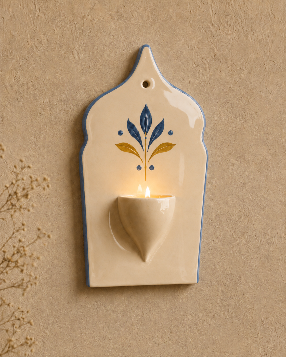 An off-white ceramic wall sconce candle holder with a blue border and a blue and gold floral motif, featuring a lit candle inside, mounted on a textured beige wall.