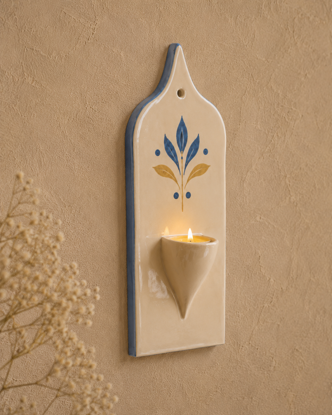 A cream-colored ceramic wall sconce candle holder with a blue and gold painted floral motif, mounted on a textured beige wall with a lit candle inside.