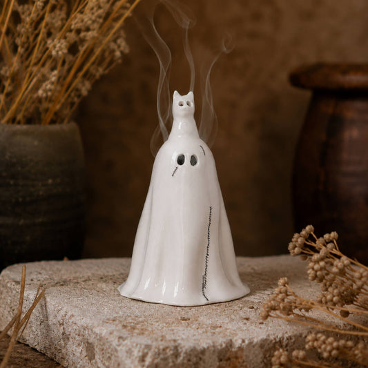A white ceramic ghost-shaped incense burner with a hooded shape, small black eyes, a stitched mouth, and smoke rising from the top, displayed on a stone slab with dried flowers and rustic pottery in the background.