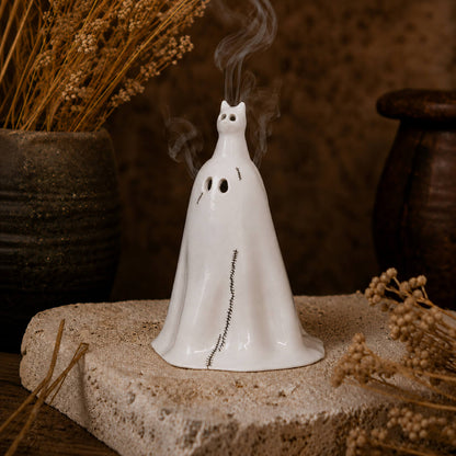 A white ceramic ghost-shaped incense holder with a cat head and small black facial details sits on a textured stone slab, with dried flowers and dark pottery in the background.
