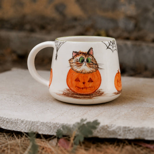 A white ceramic mug with a Halloween design, featuring a cat sitting on a jack-o'-lantern pumpkin, placed on a stone surface outdoors.