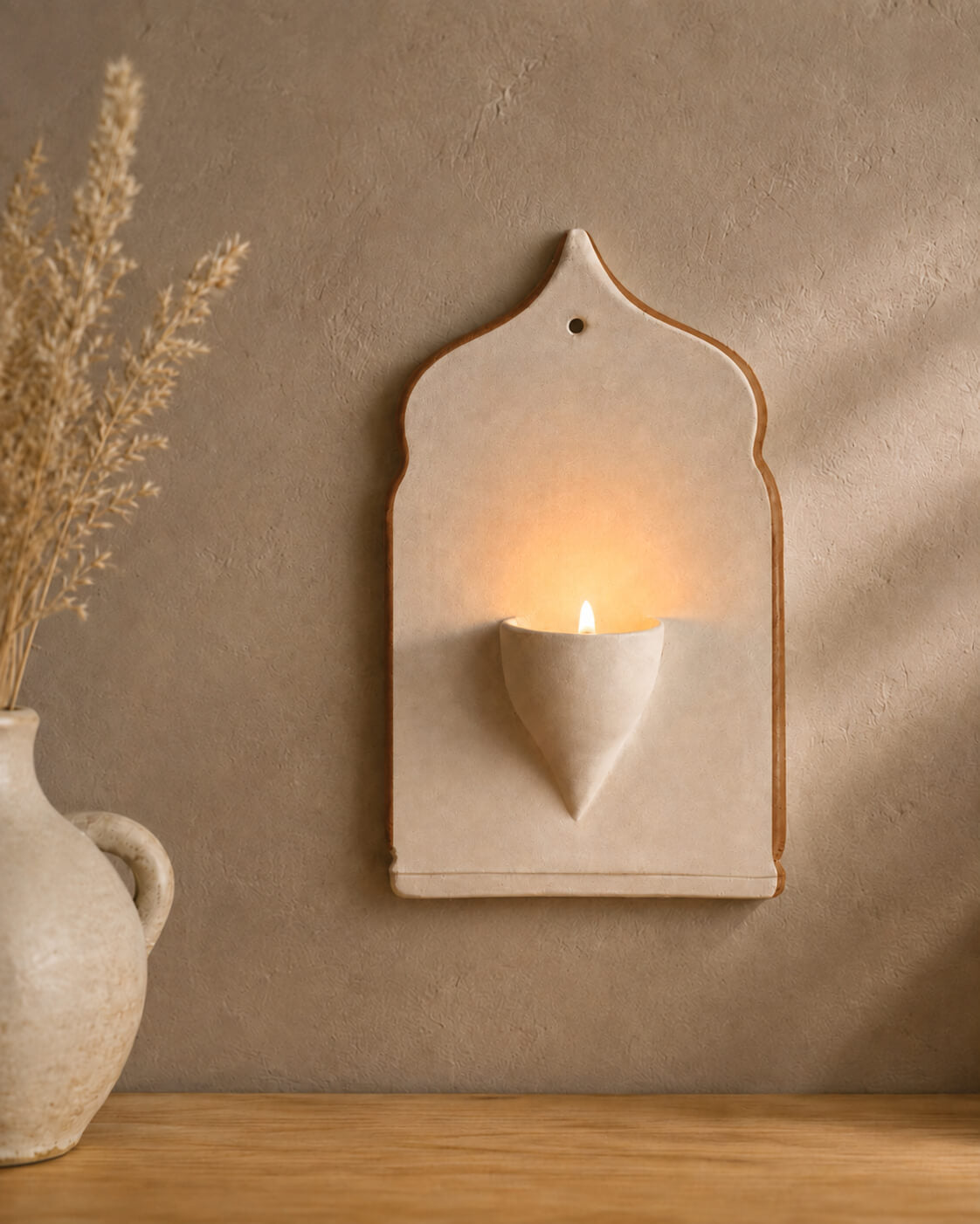 A ceramic wall sconce candle holder with a warm glowing candle, mounted on a textured beige plaster wall next to a rustic vase with dried grass.