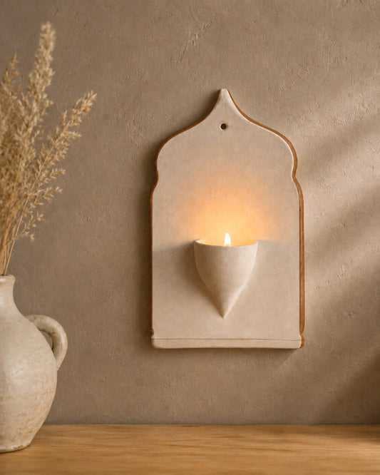 A ceramic wall sconce candle holder with a warm glowing candle, mounted on a textured beige plaster wall next to a rustic vase with dried grass.