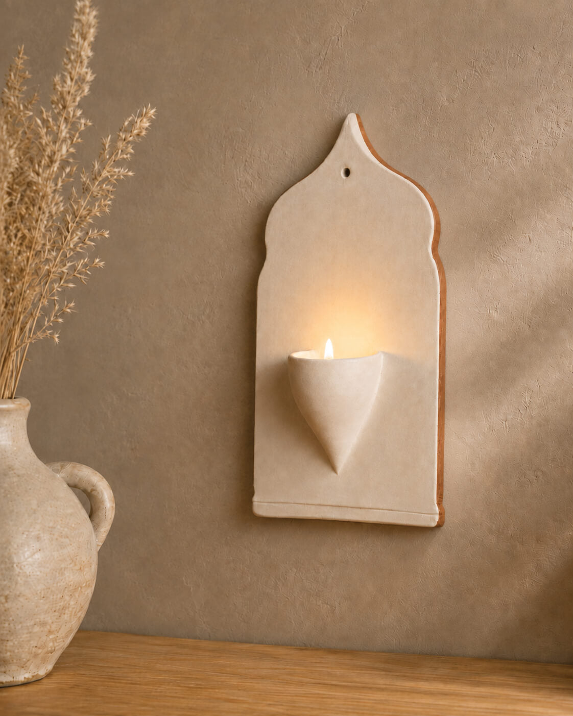 A cream-colored ceramic wall sconce holding a lit candle, mounted on a textured brown wall above a wooden surface with a ceramic vase of dried grass on the left.