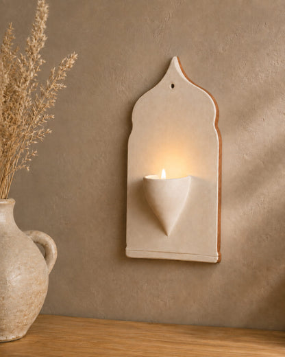 A cream-colored ceramic wall sconce holding a lit candle, mounted on a textured brown wall above a wooden surface with a ceramic vase of dried grass on the left.