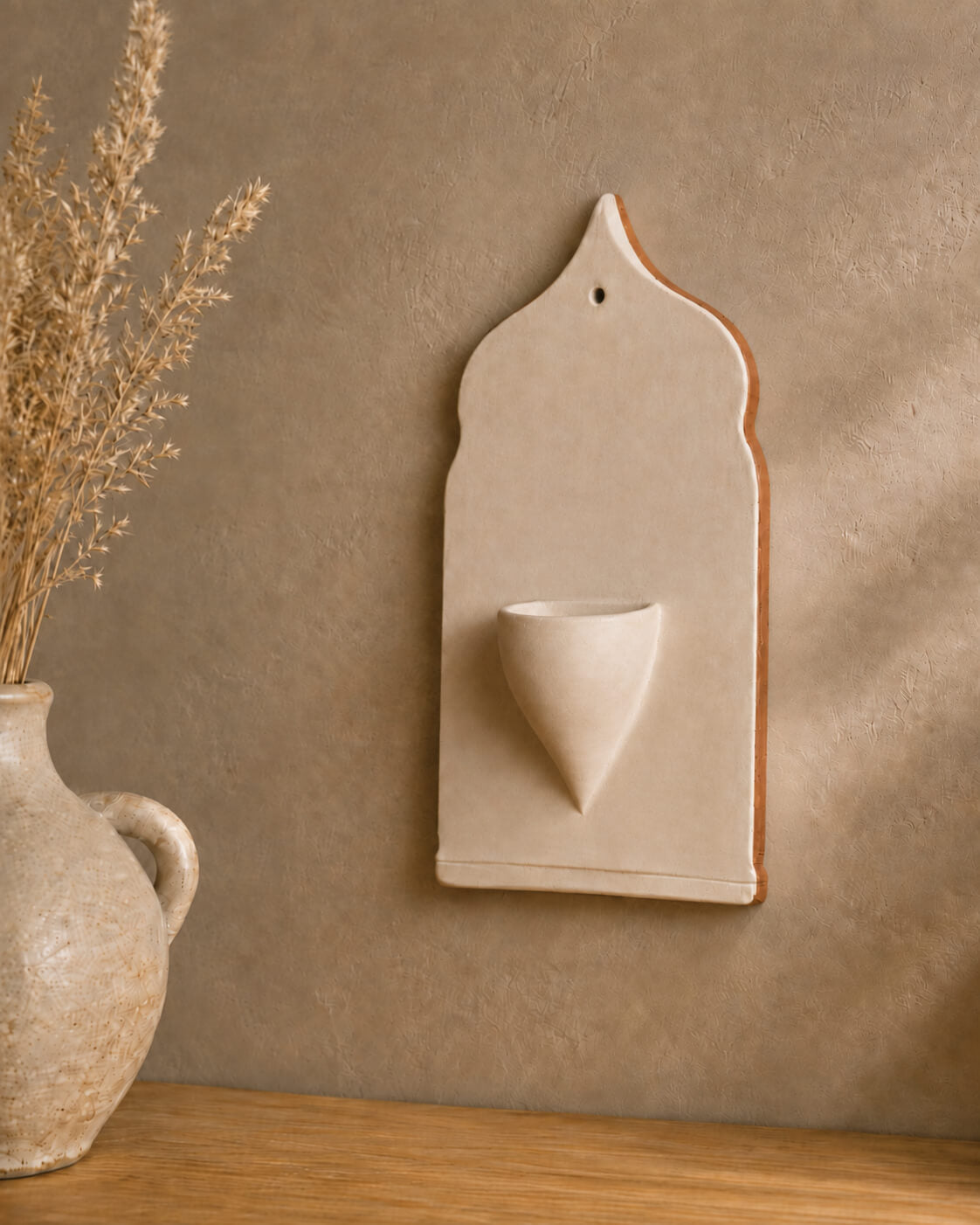 A beige ceramic wall-mounted cup holder with a small pedestal cup, styled on a wooden surface against a textured beige wall with dried grass in a vase.
