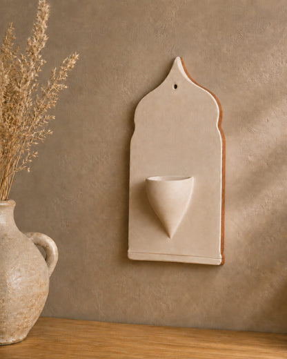 A beige ceramic wall-mounted cup holder with a small pedestal cup, styled on a wooden surface against a textured beige wall with dried grass in a vase.