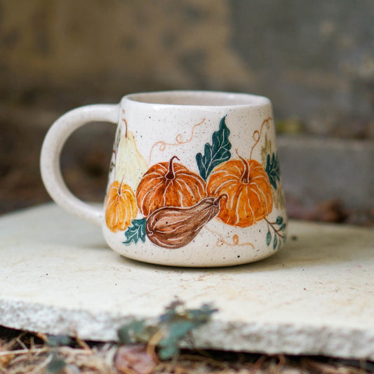 A small speckled ceramic mug with a hand-painted autumn design of orange pumpkins, a brown gourd, and green leaves, resting on a light stone slab outdoors.