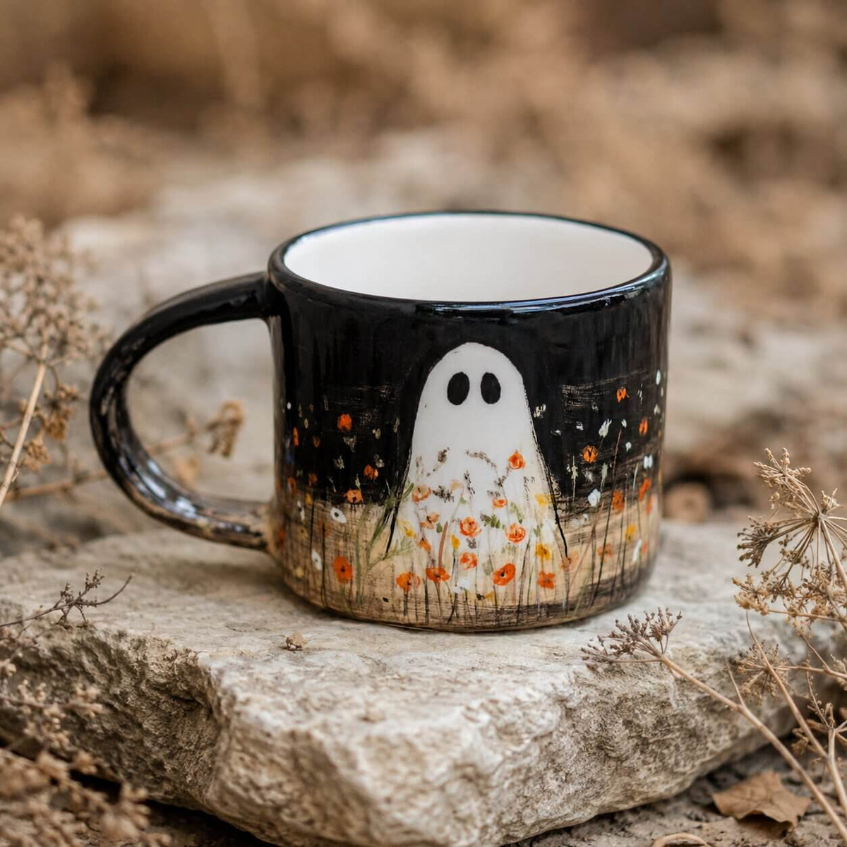 Black ceramic Halloween mug with a hand-painted white ghost and orange wildflowers, resting on a stone slab with dry grass in a natural outdoor setting.