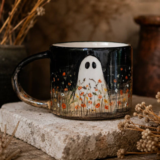 A black ceramic mug with a white ghost illustration and orange flowers, displayed on a rustic stone block with dried flowers in the background.