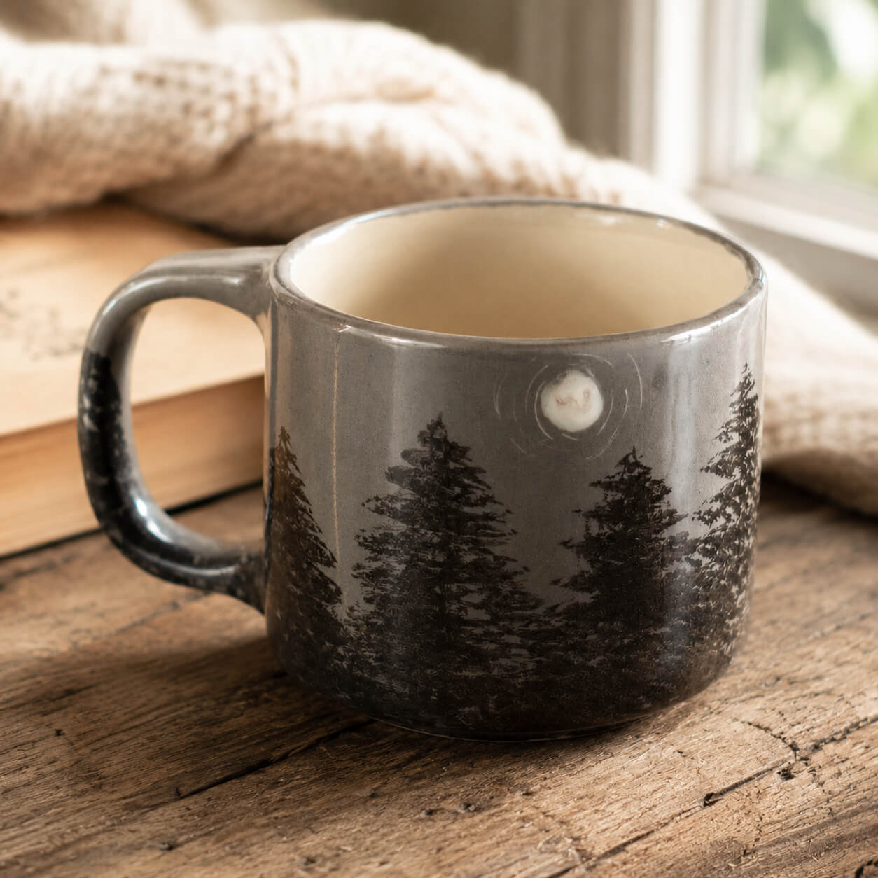 A gray ceramic mug with a black pine tree forest design, resting on a rustic wooden table with a cozy beige knit blanket and book in the background.
