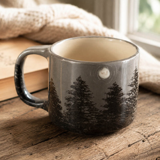 A gray ceramic mug with a black pine tree forest design, resting on a rustic wooden table with a cozy beige knit blanket and book in the background.