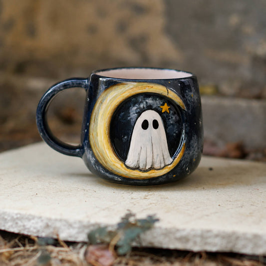 A dark ceramic mug with a Halloween design featuring a white ghost, a yellow crescent moon, and a small star, resting on a stone slab outdoors.