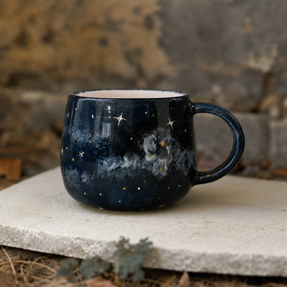 A dark blue ceramic mug with a galaxy night-sky design, featuring white stars, white nebulae, and small gold star accents, resting on a stone slab outdoors.