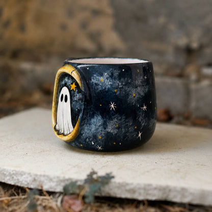 A small dark ceramic cup or candle holder with a hand-painted celestial and ghost design, resting on a stone slab outdoors.