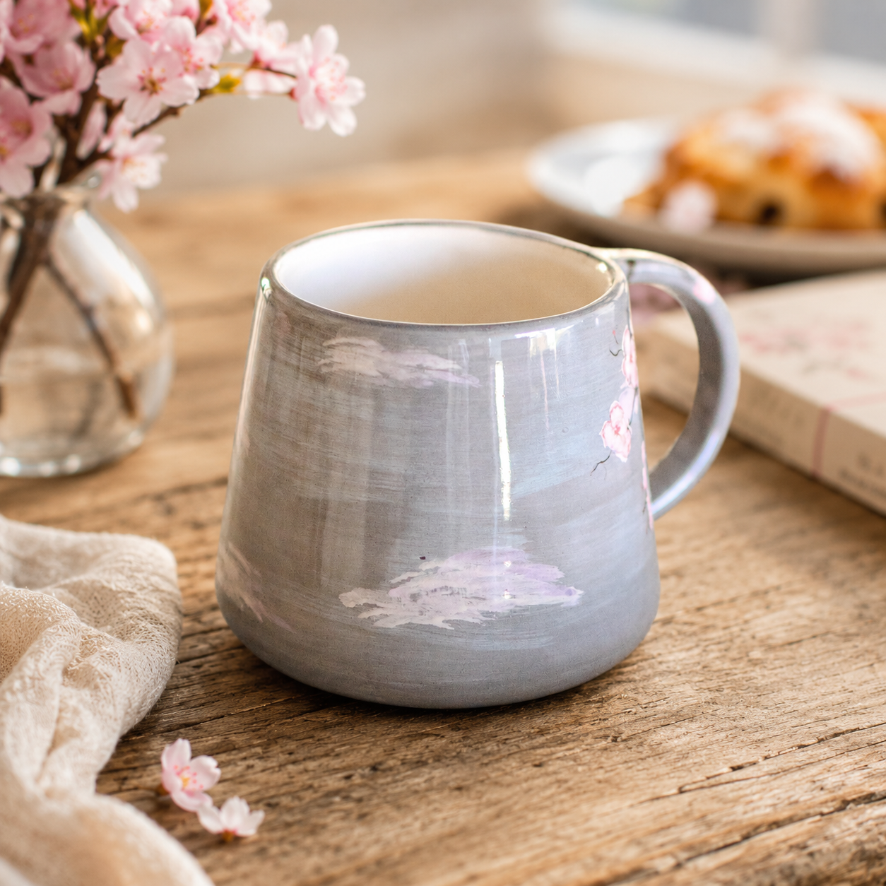 A gray ceramic mug with a hand-painted cherry blossom and cloud design, sitting on a rustic wooden table with a blurred background of flowers and a pastry.