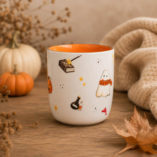 A white ceramic mug with an orange interior, decorated with cute Halloween illustrations including a ghost, pumpkin, potion bottle, and stars, set on a wooden table with autumn props.