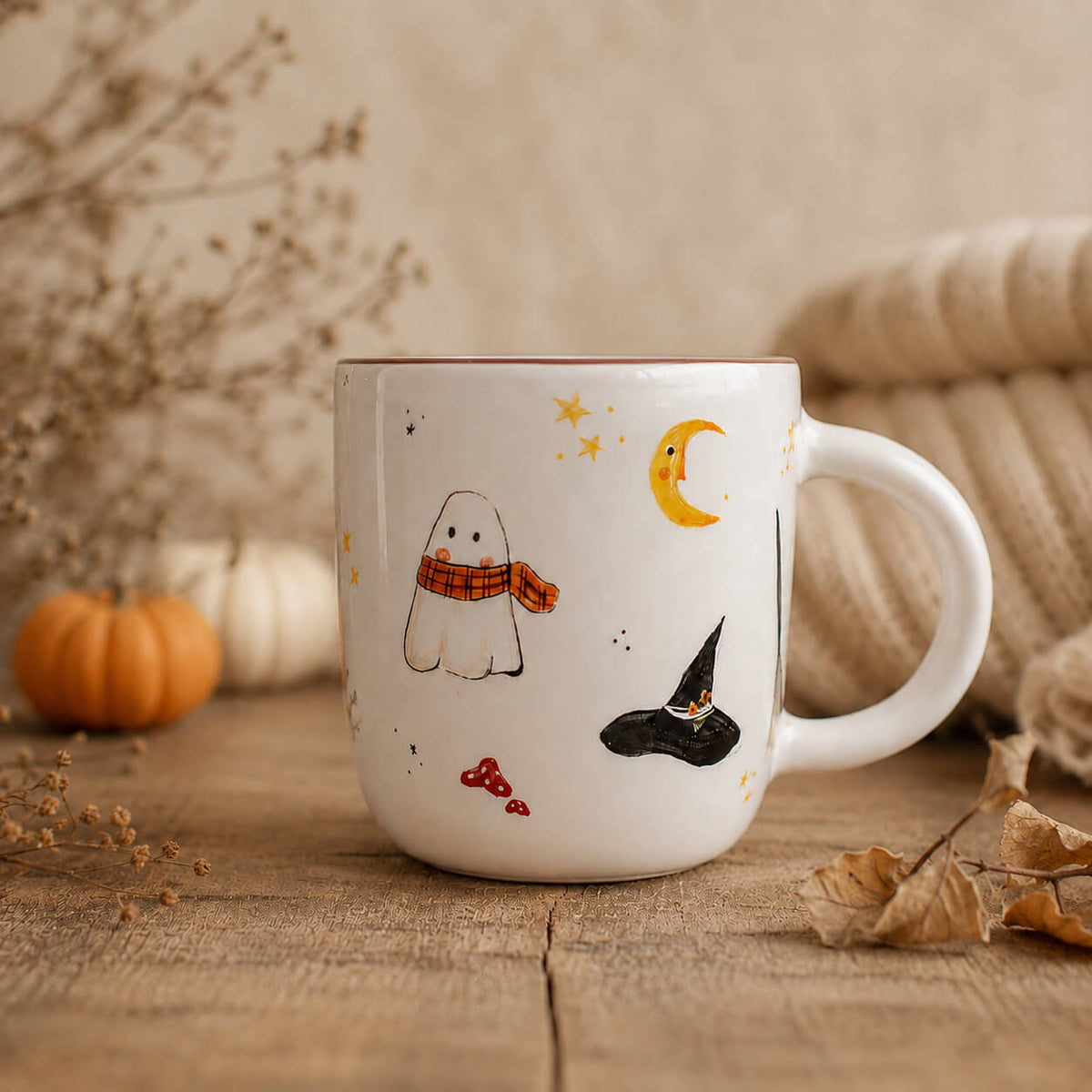 A white ceramic mug decorated with cute Halloween illustrations including a ghost, witch hat, stars, and a crescent moon, styled on a rustic wooden table with autumn props.