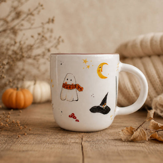 A white ceramic mug decorated with cute Halloween illustrations including a ghost, witch hat, stars, and a crescent moon, styled on a rustic wooden table with autumn props.