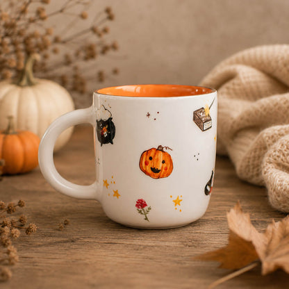 A white ceramic mug with an orange interior, decorated with a cute Halloween theme including a jack-o'-lantern, black cat, stars, and a small book. The mug is set on a rustic wooden table surrounded by autumn props like a burlap sack, dried flowers, and small pumpkins.