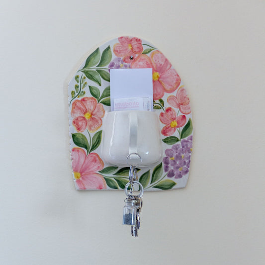 Hand-painted ceramic key holder adorned with colorful flowers on a white wall. Handbemalter keramischer Schlüsselhalter, verziert mit bunten Blumen an einer weißen Wand.