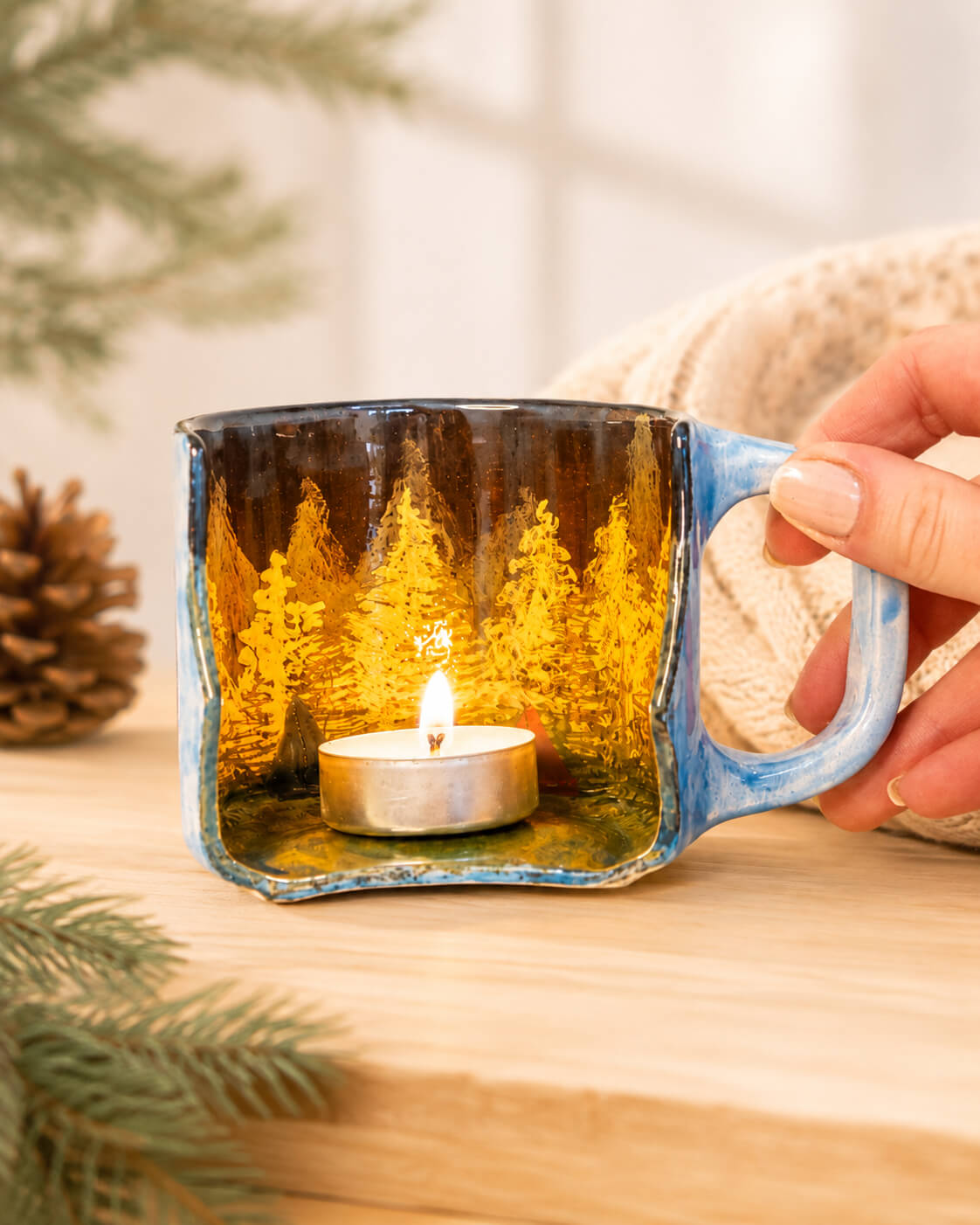 A hand holds a square ceramic mug with a glowing tealight candle inside, set against a warm, cozy background with pine branches and a knitted blanket.