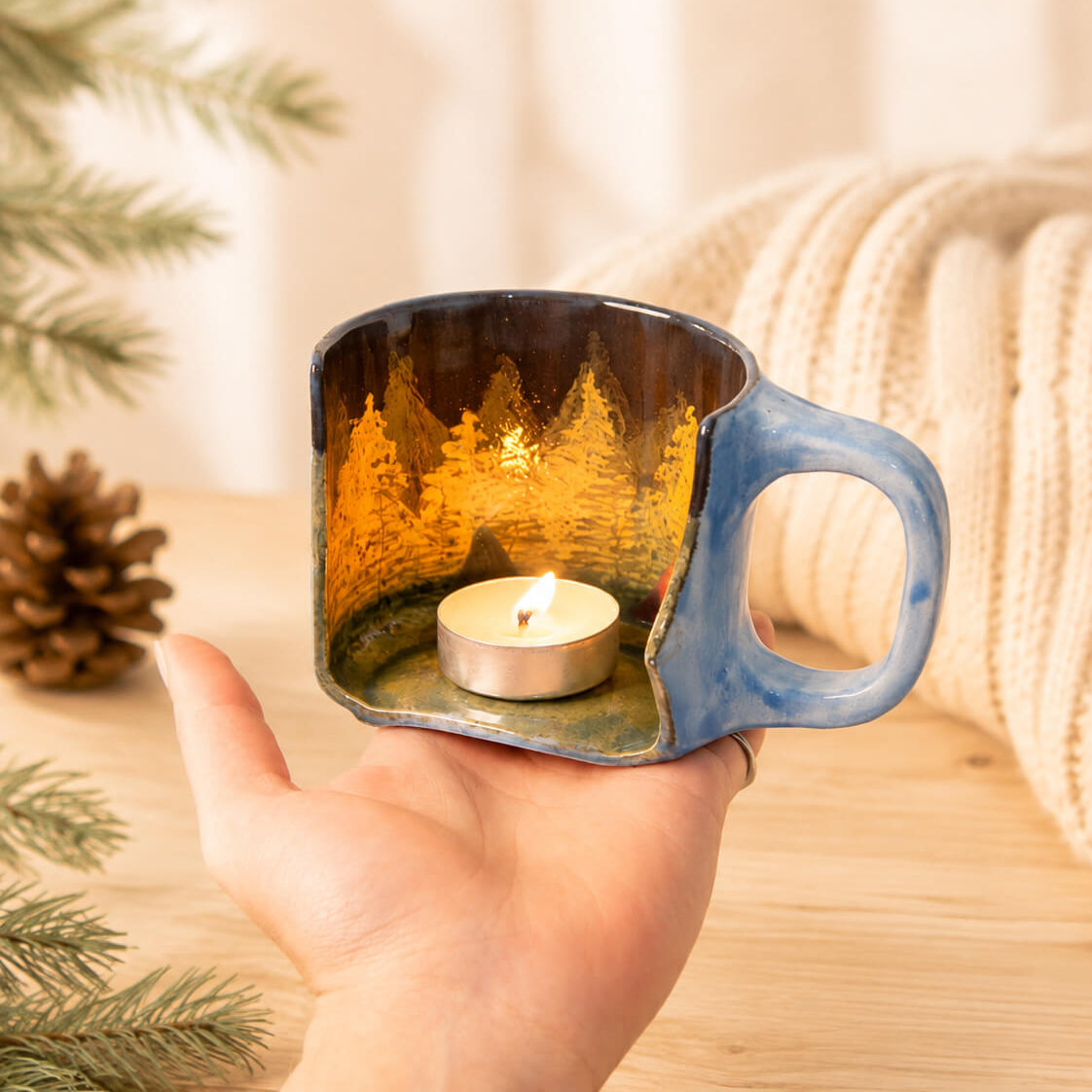 A hand holds a small square ceramic candle holder with a blue-handled mug design and a warm forest scene printed inside. A tea light candle burns inside, and pine branches, a pinecone, and a cream knit blanket appear softly blurred in the background.