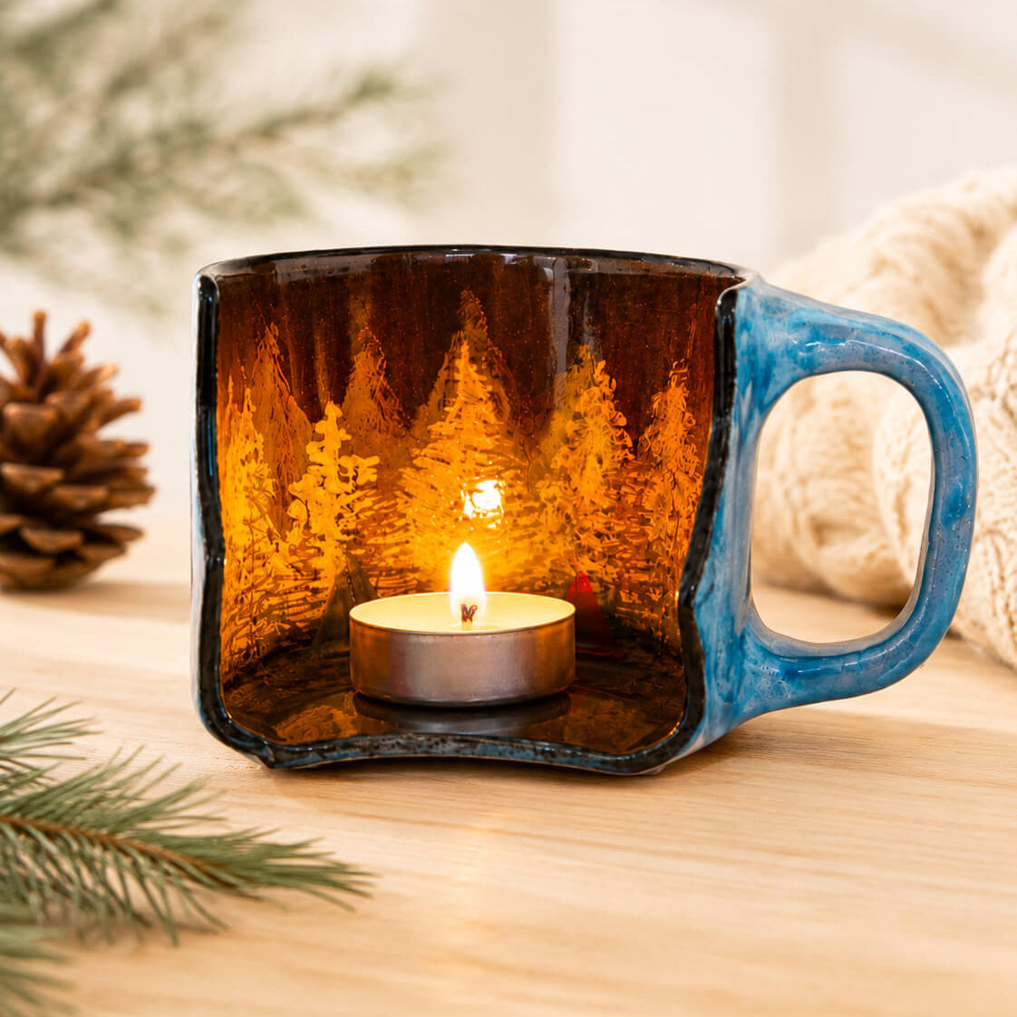 A square ceramic mug candle holder with a blue handle and a forest scene printed inside, holding a lit tealight candle on a wooden surface with pine branches and a knitted blanket in the background.
