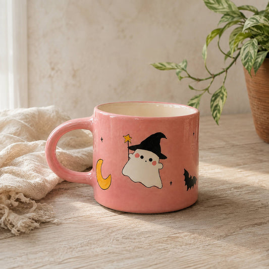 A pink ceramic mug with a cute Halloween design featuring a white ghost in a black witch hat, a yellow crescent moon, and a black bat, set on a wooden table next to a beige throw blanket and a potted plant.