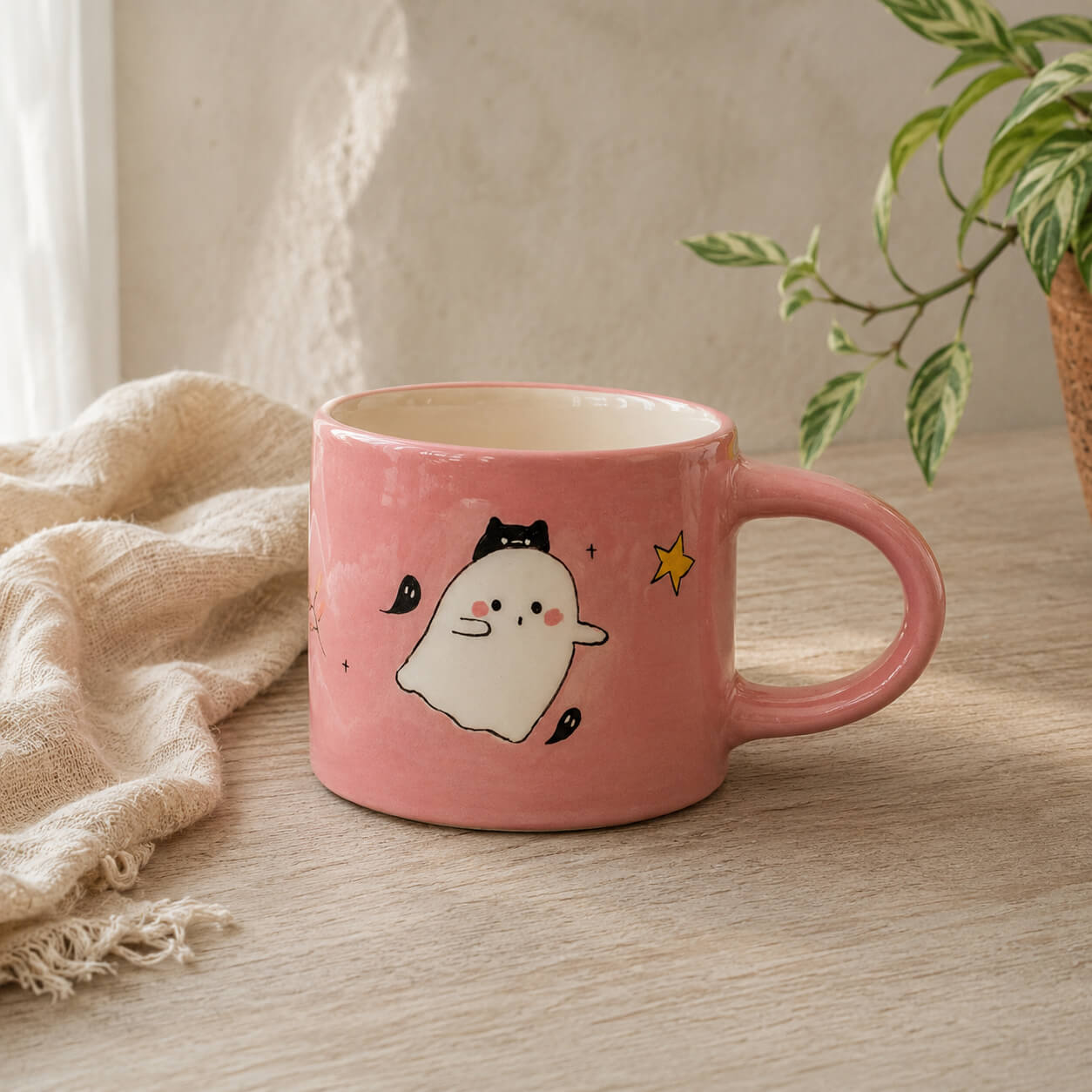 A pink ceramic mug with a cute cartoon ghost character and a small yellow star, styled on a wooden table with a beige cloth and a potted plant in the background.