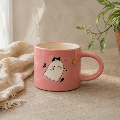 A pink ceramic mug with a cute cartoon ghost character and a small yellow star, styled on a wooden table with a beige cloth and a potted plant in the background.