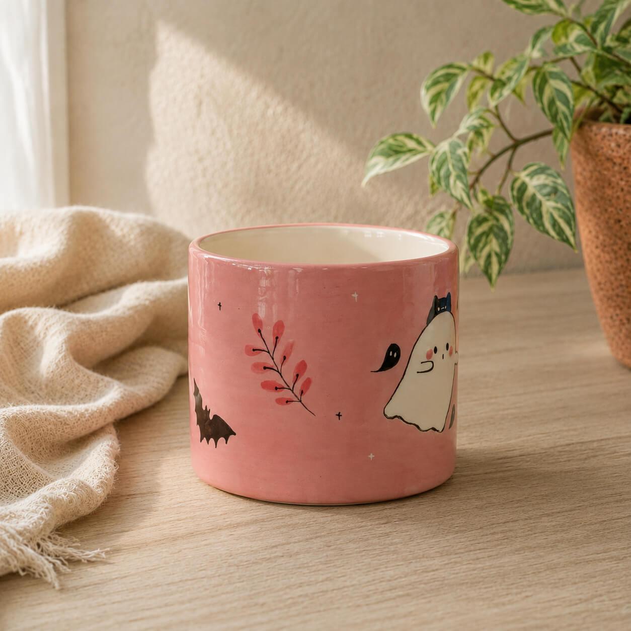 A pink ceramic mug with cute Halloween illustrations, including a ghost, bat, and red berries, sitting on a wooden table next to a beige throw blanket and a potted plant.