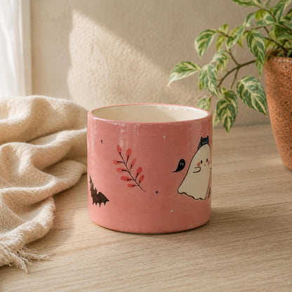 A pink ceramic mug with cute Halloween illustrations, including a ghost, bat, and red berries, sitting on a wooden table next to a beige throw blanket and a potted plant.