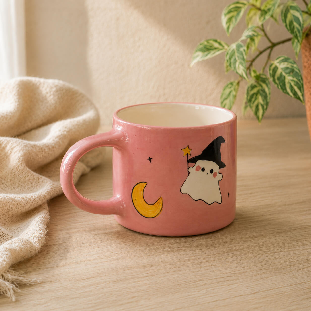 A pink ceramic mug with a cute cartoon ghost and crescent moon design, sitting on a wooden table next to a beige knit blanket and a green plant.